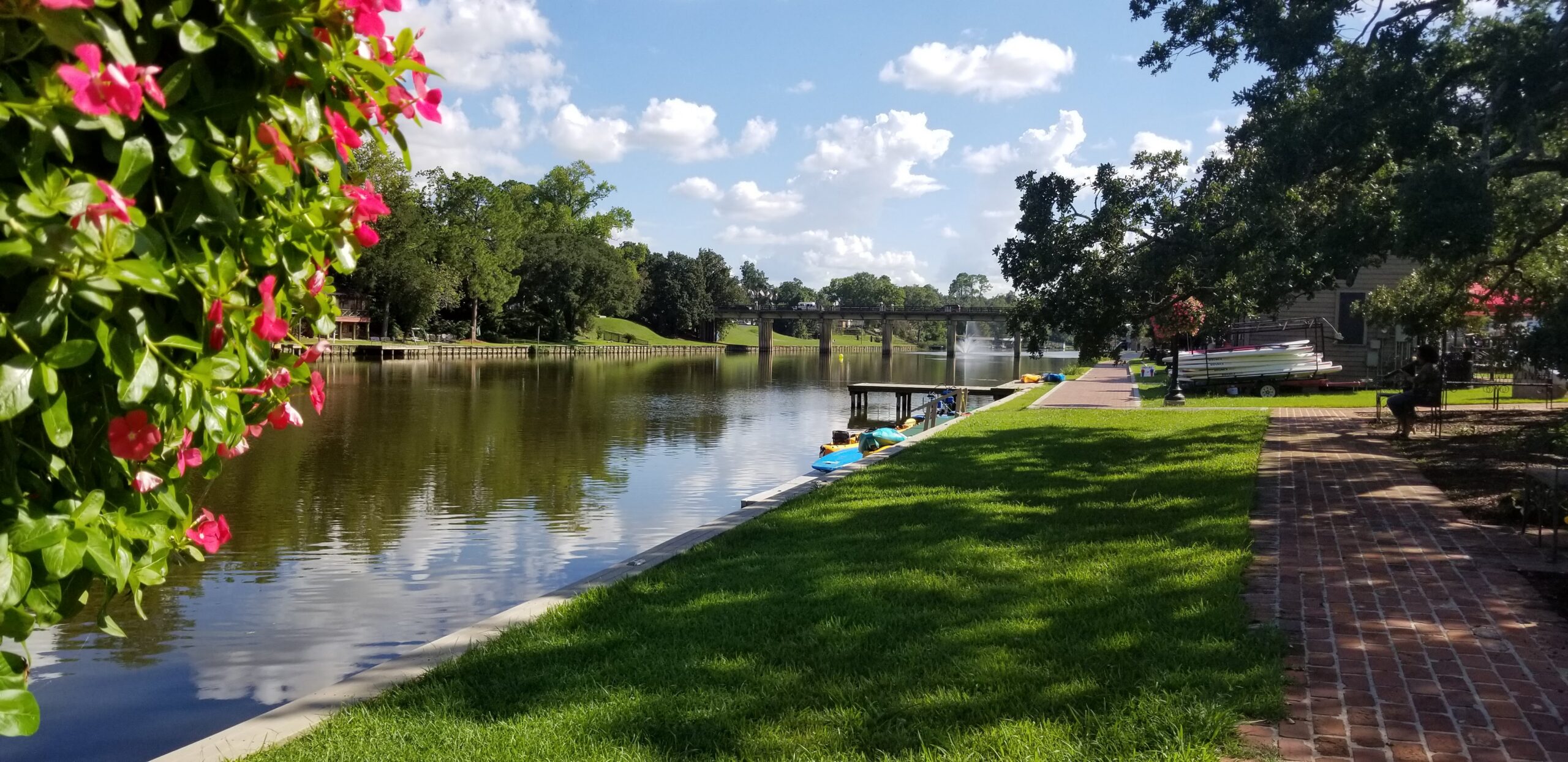 Tranquil Cane River park scene with pond, brick walkway, trees, fountain, and pink flowers