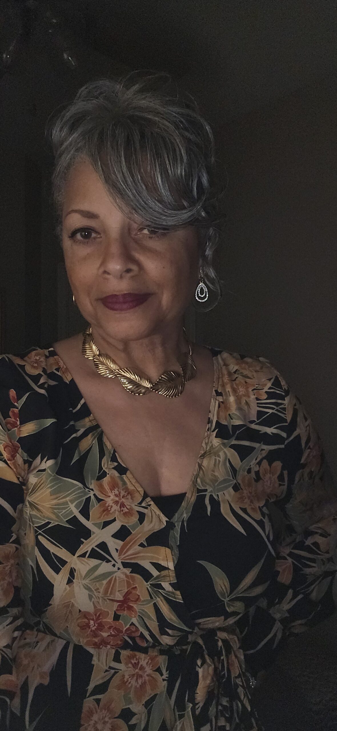 Portrait of a woman with silver hair, floral top, and gold jewelry looking at the camera